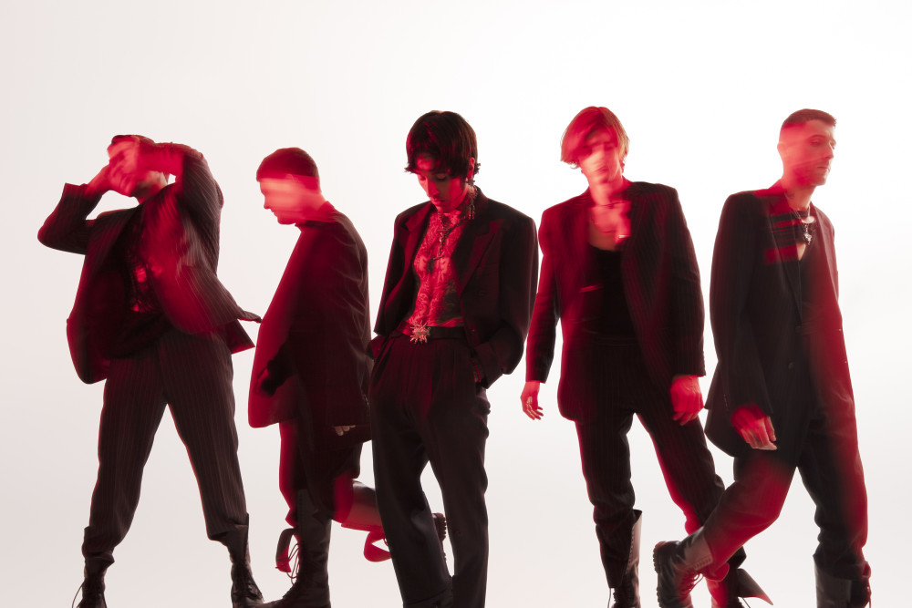Bandfoto von Bring Me The Horizon: Fünf männlich gelesene Personen in schwarzen Anzügen stehen vor einer weißen Wand. Sie sind mit einem roten Filter-Verwisch-Effekt versehen, sodass nur das Gesicht der in der Mitte stehenden Person erkennbar ist.
