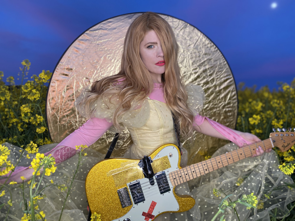 Eine weiblich gelesene Person mit langem, blonden Haar und golgenem Tüll-Kleid steht inmitten eines Rapsfeldes und hat eine gold-glitzerne Gitarre umgehängt. Hinter ihr wird eine goldene Scheibe gehalten.