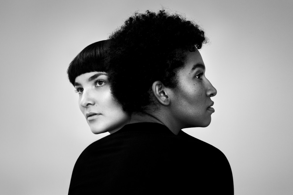 Schwarz/weiß Fotografie der Band Olicía: Zwei weiblich gelesene Personen schmiegen sich aneinander, die Gesichter sind voneinander abgewandt.