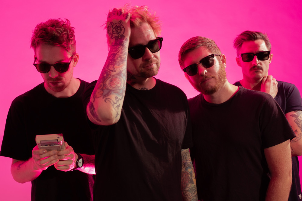 Vier männlich gelesene Personen mit schwarzen T-Shirts und Sonnenbrillen vor einem neon pink strahlendem Hintergrund.