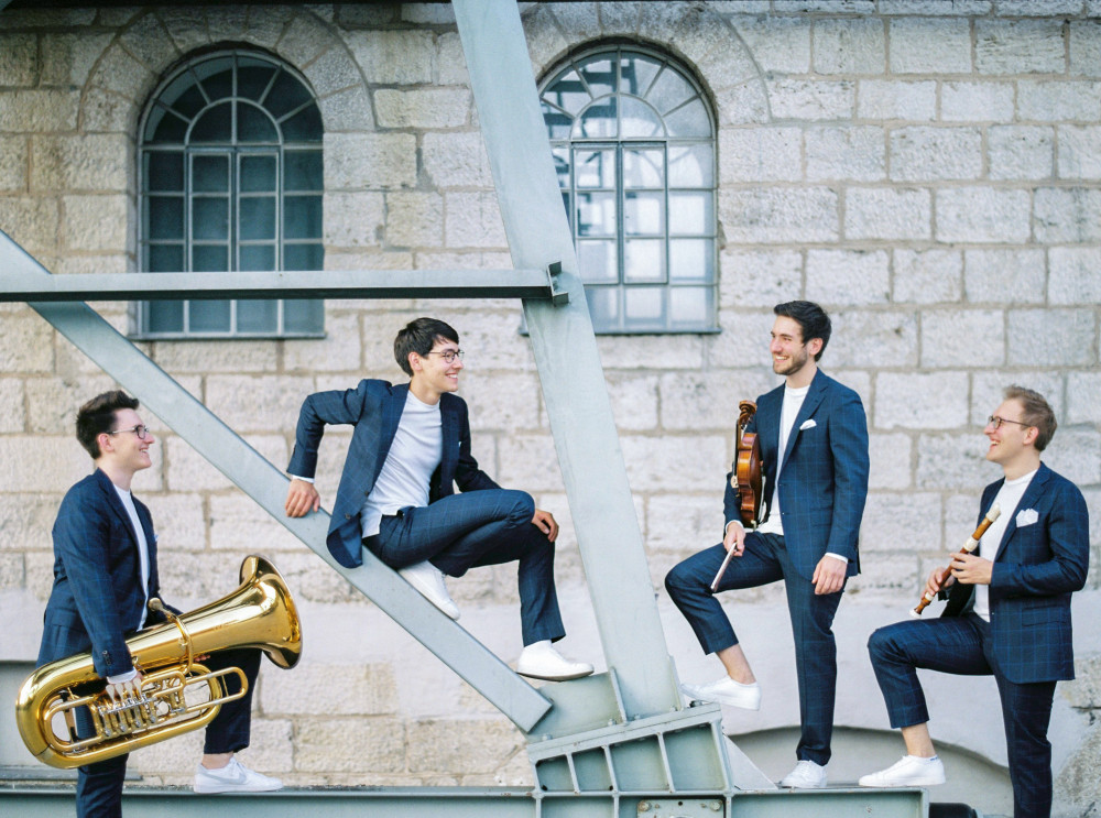 Gruppenfoto der Hanke Brothers: Vier männlich gelesene Personen in den gleichen blauen Anzügen stehen vor einer Steinwand und stehen/sitzen auf einem Stahlträger.