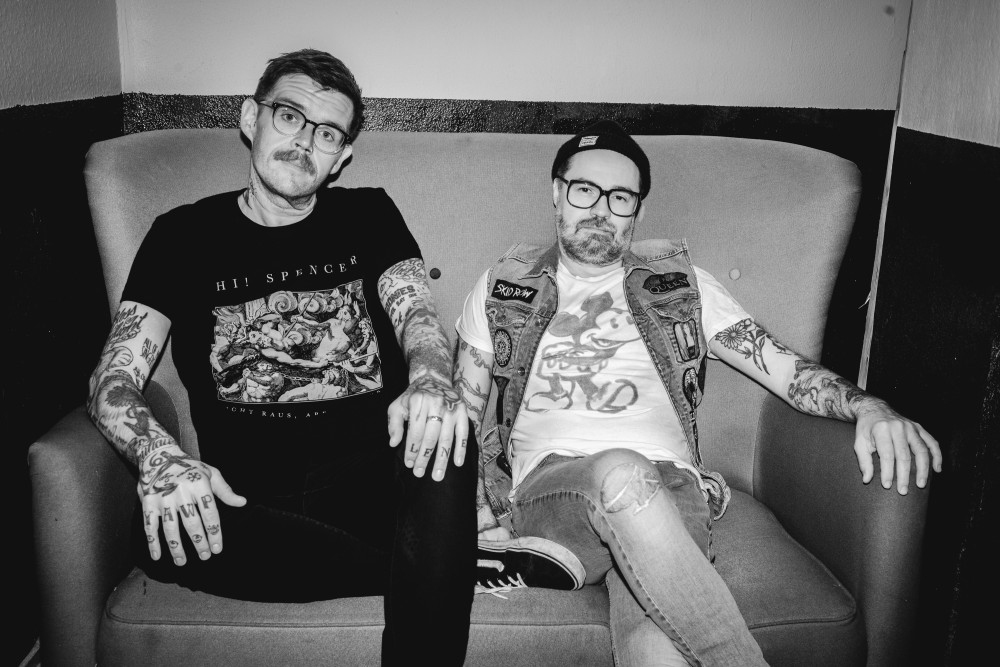 Schwarz/weiß Fotografie der Band Jupiter Jones: zwei männlich gelesene Personen sitzen auf einer Couch vor einer Wand und blicken in die Kamera. Beide tragen eine Brille.