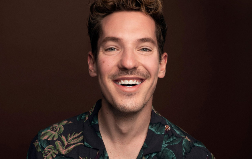 Es ist eine lachende, männlich gelesene Person vor einem hellgrauen Hintergrund zu sehen. Die Person trägt einen bedruckten schwarzen Anzug. Auf dem Anzug sind Sonnen Symbole sowie Smileys und Sonnenblumen aufgedruckt. Die Person sitzt auf einem weißen Hocker. Die Person hat kurze, dunkle Haare. Im Hintergrund ist der weiße Schriftzug "NICO STANK" zu lesen, die Person verdeckt einen Teil des Schriftzugs. Im unteren Bildbereich ist der orangene Schriftzug "merkste selber" zu lesen.