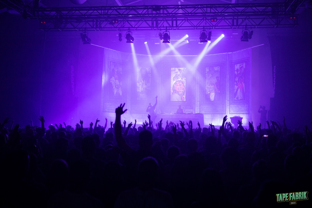 Konzertfotografie aus der Halle des Kulturzentrums Schlachthof bei der Tapefabrik 2023: Eine Menschenmasse mit in die Höhe gestreckter Hände steht vor einer Bühne, auf der ein männlich gelesener Künstler rappt.