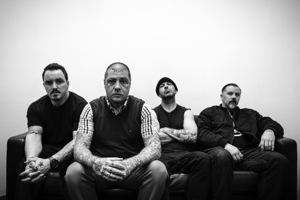 Schwarz/weiß Fotografie der Band Rancid. Vier männlich gelesene Personen sitzen nebeneinander vor einer weißen Wand auf einer Couch und blicken mit ernstem Blick in die Kamera.