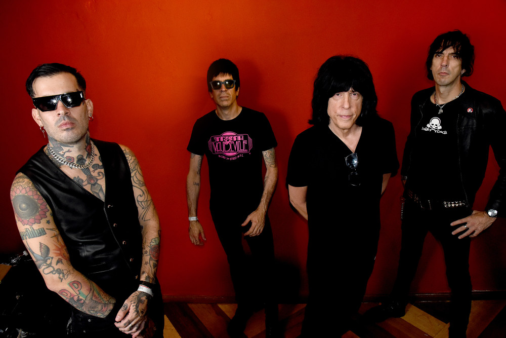 Bandfoto von Marky Ramone: Vier männlich gelesene Personen stehen vor einer roten Wand, alle sind schwarz gekleidet. Der zweite von rechts ist Marky Ramone. Er trägt langes schwarzes Haar.
