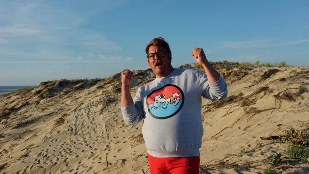 Es ist eine männlich gelesene Person vor einer Dünenlandschaft zu sehen. Die Person hat die Fäuste gereckt und den Mund geöffnet. Die Person trägt einen graues, bedrucktes Sweatshirt mit einem Snoopy Motiv. Die Person trägt eine Brille und hat einen Schnurrbart sowie mittellanges, braunes Haar.