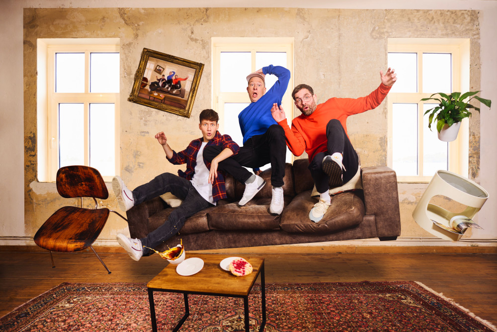 Bandfoto von Deine Freunde: Drei männlich gelesene Personen schweben auf einer Couch sitzend in der Luft und schauen erstaunt. Die Gegenstände um sie herum schweben ebenfalls in der Luft, als hätte man sie fallen lassen