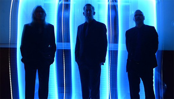 Drei männlich gelesene Körper stehen vor blauem Hintergrundlicht und sind schemenhaft zu erkennen
