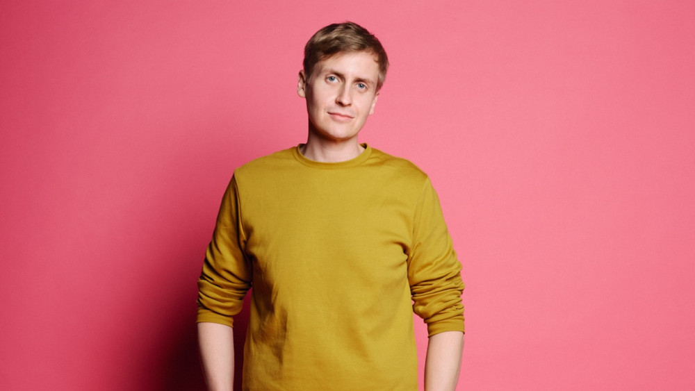 Zu sehen ist eine männlich gelesene Person in einem senfgelben Pullover, der vor einem rosa farbenen Hintergrund steht.