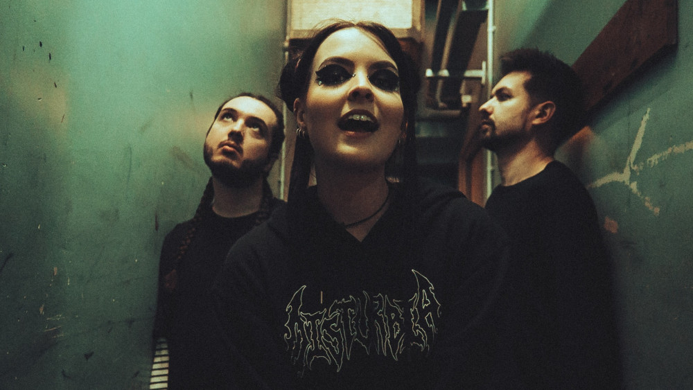 Zu sehen sind drei dunkel gekleidete Personen, die in einem Gang stehen. Während die mittlere zum Betrachteter schaut, blicken die anderen zwei Personen in verschiedene Richtungen.