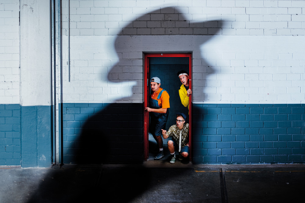 Drei bunt gekleidete, männlich gelesene Personen schauen gespannt durch eine Türöffnung nach links, von wo sich ein dunkler, menschlicher Schatten mit Hut über die Szenerie zieht.