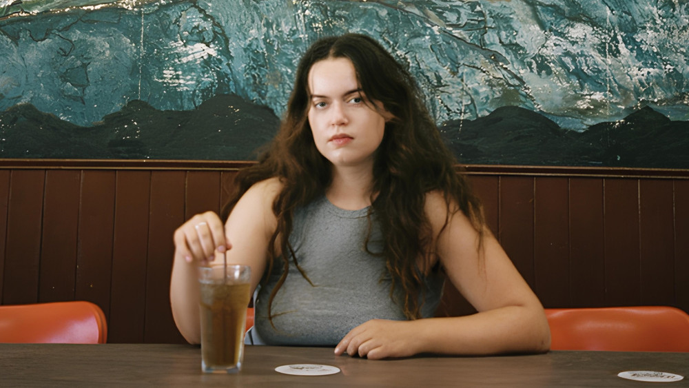Zu sehen ist eine weiblich gelesene Person im Porträt, die an einem langen Tisch sitzt. Während ihr linker Arm locker darauf aufliegt, rührt sie mit der rechten Hand in einem Getränk und schaut den Betrachter an.