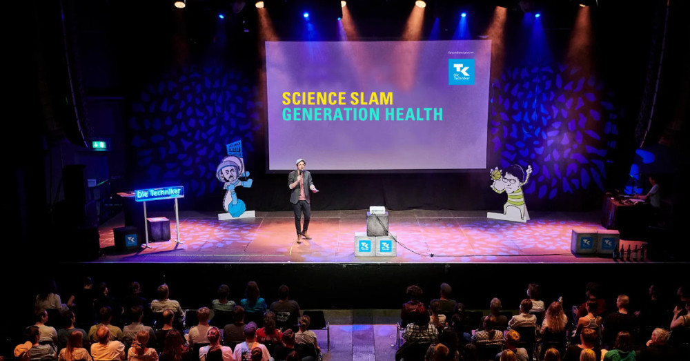 Zu sehen ist eine Bühne, auf der eine moderierende Person steht. Hinter ihr sind blaue Lichter und eine Leinwand mit der Aufschrift "Science Slam Generation Health" zu sehen.
Vor der Bühne sitzt das Publikum.