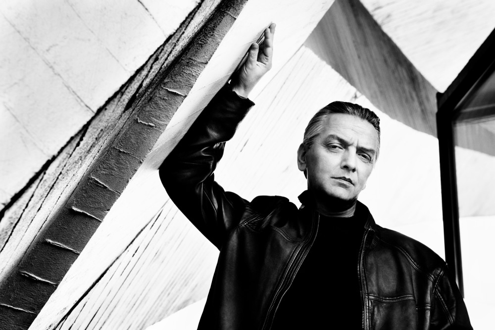 Schwarz/weiß Fotografie des Künstlers* Peter Heppner, der eine Lederjacke trägt und sich mit einem Arm an eine schräge Wand stützt. Sein Blick ist zum Betrachter gerichtet.