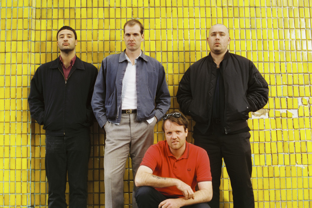 Vor einer gelben Fliesenwand stehen drei männlich gelesene Personen in dunkler Kleidung. Eine vierte Person in einem roten Oberteil kniet davor. Alle vier haben einen neutralen Gesichtsausdruck und schauen den Betrachter an.