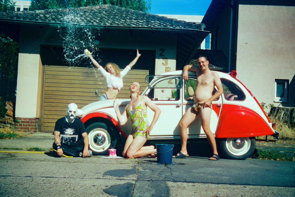 Bandfoto von Mehnersmoos: vier leicht bekleidete Personen stehen um einen rot-weißen Oldtimer "Ente" herum und waschen ihn mit viel Hingabe.