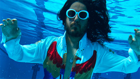 Zu sehen ist eine männlich gelesene Person unter Wasser in einem Schwimmbecken. Sie hat eine Sonnenbrille auf  und lange dunkle Haare. Um den Hals hängt eine Kette.