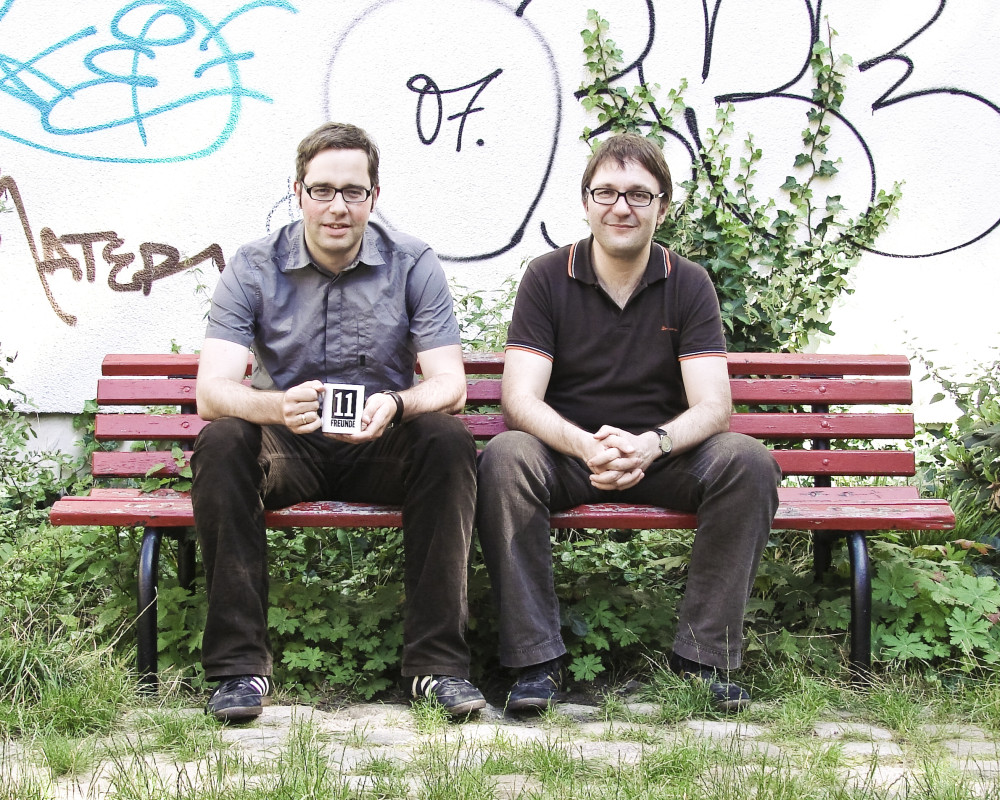 Zwei männlich gelesene Personen mit Brille sitzen auf einer roten Holzbank vor einer Betonmauer und blicken in die Kamera. Die links sitzende hält eine Tasse, auf der "11 Freunde" steht in den Händen.