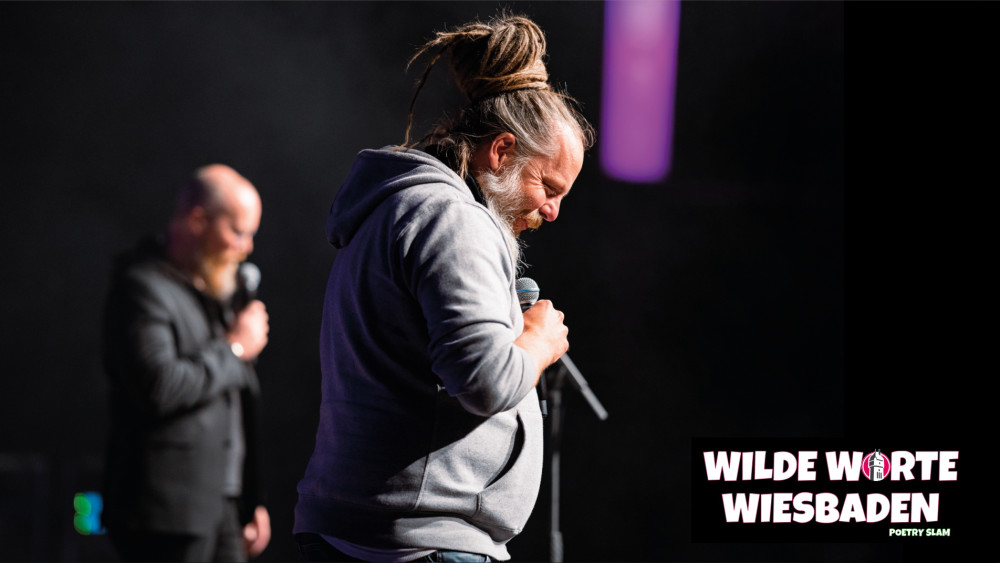 Es ist eine männlich gelesene Personen vor einem dunklen Hintergrund zu sehen. Sie trägt einen grauen Kapuzenpullover, einen grauen Bart und zu einem Dutt geflochtene Dreadlocks. In der rechten unteren Ecke des Bildes ist der weißer Schriftzug "WILDE WORTE WIESBADEN" zu lesen.