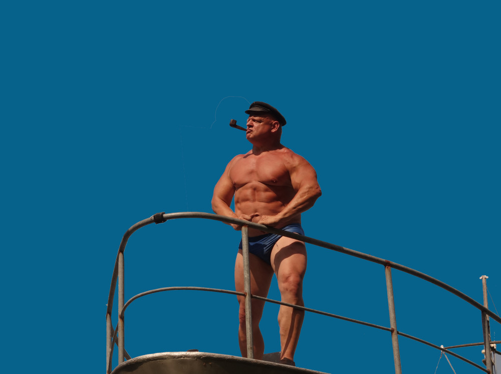 Eine sehr muskulöse, männlich gelesene Person mit Batschkapp auf dem Kopf und Pfeife im Mund steht mit einer Badehose bekleidet an einem Geländer vor blauem Hintergrund.