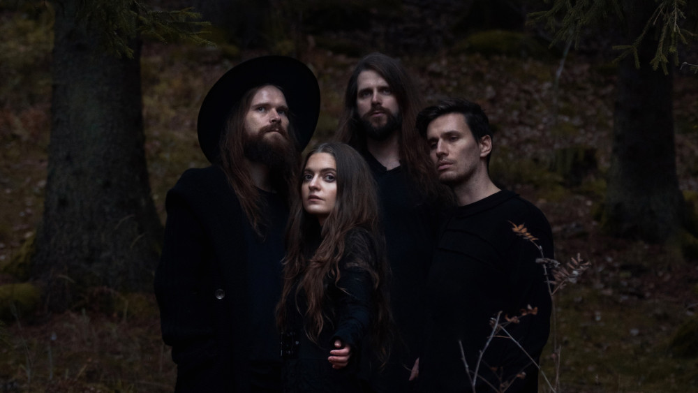 Auf dem Foto sieht man 3 männlich gelesene und eine weiblich gelesene Person. Das Bild ist sehr dunkel und in einem Wald. Die Person ganz rechts hat lange braune haare mit einem Langen Bart.Er trägt eine schwarzen großen Hut und eine Schwarze Jacke.
Rechts vor ihm eine Frau mit langen braunen Haaren und einer schwarzen Jacke. Hinter ihr auch eine Person mit langen Braunen Haaren und schwerem Oberteil. Links von ihm vorne dran ist eine Person mit Braunen Haaren und einem Schwarzen Oberteil. Sie gucken alle in die Kamera.