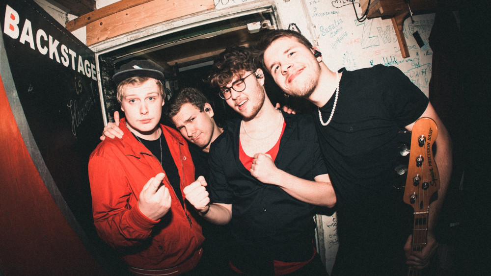 Das Foto auf dem vier Männlich gelesene Personen zu erkennen sind ist vor einer offenen Backstage Tür. Die Person ganz Rechts hat kurze Braune Haare und einen drei Tage Bart. Er trägt ein schwarzes Shirt und hat eine Perlenkette um. Er hält auch seinen Gitarrenkopf in der Hand. Die Person rechts daneben hat Braune kurze lockige Haare und ein weißes kurzes Hemd mit weitem Ausschnitt wodurch man noch sein rotes Shirt erkennt. Er hat außerdem eine schwarze Brille. Daneben stet eine Person mit Cap wodurch blonde haare zum Vorschein kommen. Dazu trägt er eine Rote Jacke mit einem Schwarzen Shirt drunter. Hinter den beiden letzten beschriebenen Personen sieht man noch einen Kopf zwischen den Schultern mit kurzen lockigen Haaren.