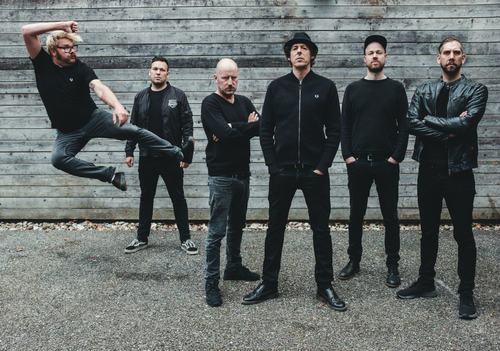 Auf dem Bild sind sechs männlich gelesene Personen vor einer Holzwand auf einer Wiese zu sehen. Die Umgebung wirkt sehr grau. Die Person links im Bild springt in die Luft und trägt kurze, blonde Haare, einen braunen Bart und eine Brille. Außerdem trägt sie eine graue Jeans, ein schwarzes T-Shirt und schwarze Schuhe. Die Person rechts neben ihr trägt kurze, braune, nach oben gestylte Haare und eine schwarze Hose, ein schwarzes T-Shirt mit einer schwarzen Jacke mit weißem Aufdruck und schwarz/weiße Sneaker. Die Person rechts von ihr hat die Arme verschränkt, trägt eine Glatze, einen Bart, graue Jeans, ein schwarzes, langärmliges Oberteil und Schwarze Schuhe. Die Person rechts von ihr trägt dunkle, mittellange Haar und einen schwarzen Hut darüber. Sie ist ebenfalls schwarz mit einer Hose, einer geschlossenen Jacke und Schuhen bekleidet. Die Person rechts von ihr hat rote Haare, die von einer Kappe verdeckt sind und trägt einen Bart. Sie trägt ebenfalls schwarze Kleidung, genauer gesagt schwarze Schuhe, eine schwarze Hose, einen schwarzen Gürtel sowie ein langärmliges schwarzes Oberteil. Die Person rechts von ihr trägt kurze braune Haare und einen Bart. Sie ist ebenfalls schwarz bekleidet mit Schuhen, einer Hose, einem Oberteil und einer ebenfalls schwarzen, glänzenden Lederjacke. Bis auf die springende Person blicken alle ernst in die Kamera.