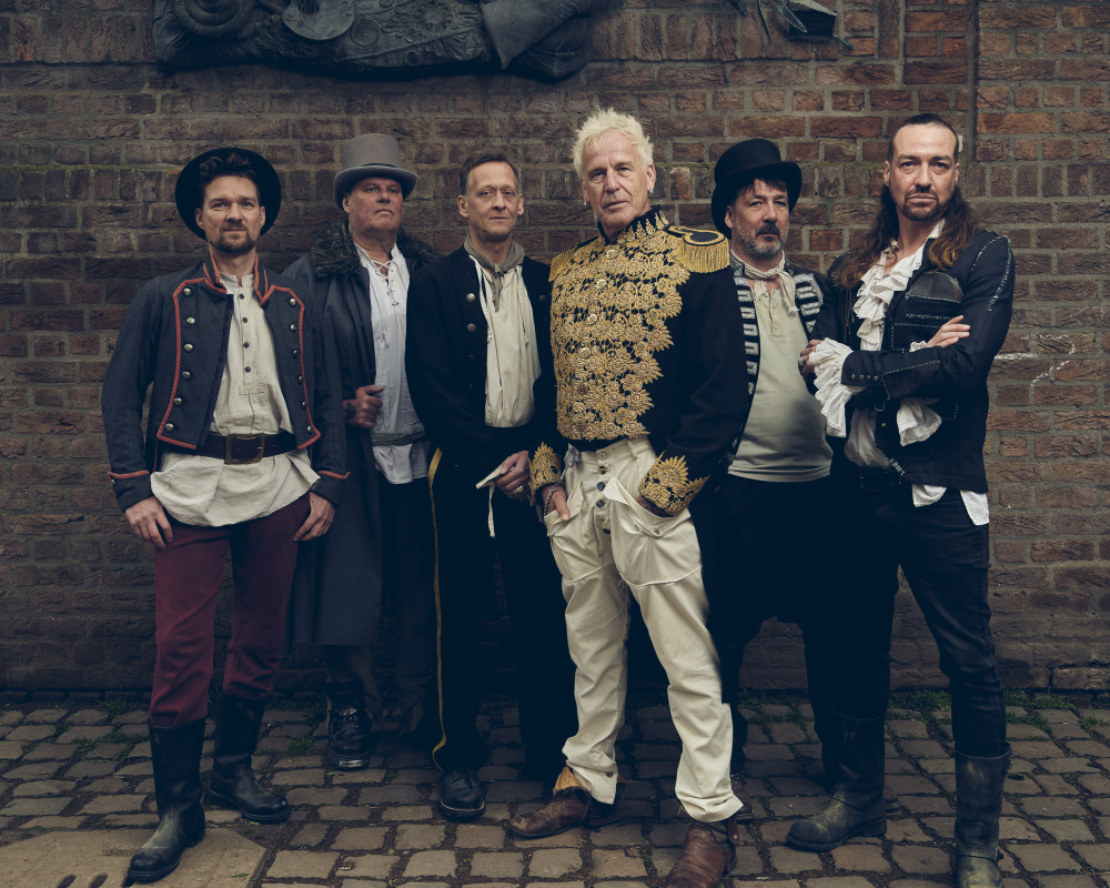 Auf dem Bild sieht man sechs männlich gelesene Personen vor einer Steinwand. Die Person ganz links trägt einen Hut, ein Hemd, darüber eine Veste. Die Person neben dran trägt eine grauen Hut ein Hemd und eine langen Mantel. Die nächste Person trägt auch ein Hemd und eine art Jackett. Die nächste Person trägt eine Jacke mit goldenen Verzierungen vorne und auf der Schulter. Die Person ganz rechts trägt ein mittelalterliches Hemd und darüber eine schwarze Jacke. Allgemein sind alle mittelalterlich gekleidet