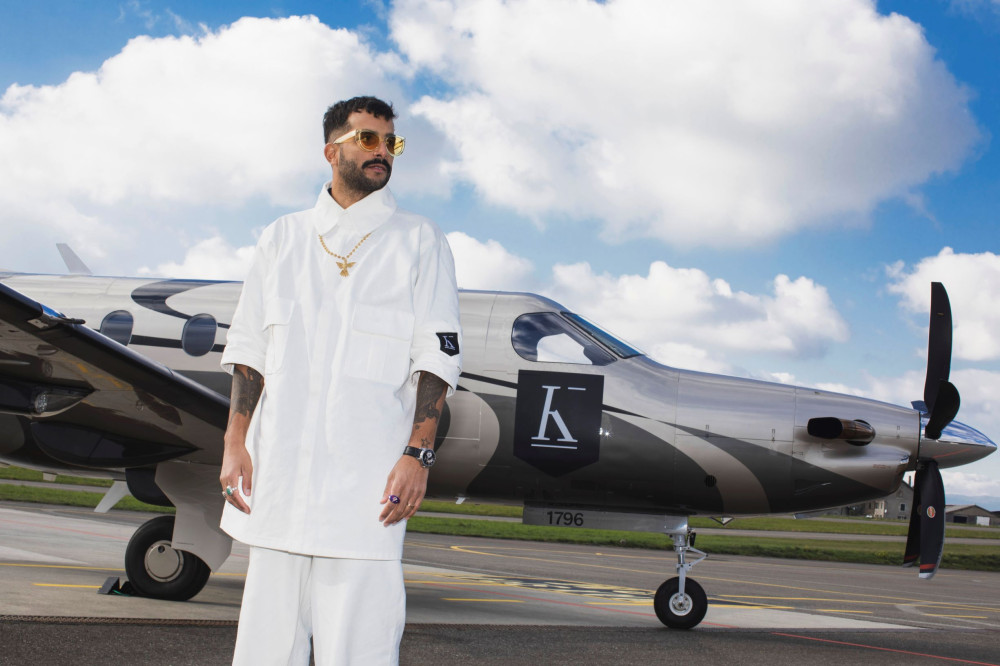 Auf dem Bild ist ein männlich gelesene Person auf eine Rollfeld vor einem Jet Flugzeug zu sehen. Die Person trägt eine Art weites weißes Hemd und Hose, eine kette und eine Sonnenbrille. Auf der Seite des Jet Flugzeugs ist eine K abgebildet. Dieses findet sich ebenfalls auf dem linken Hemdärmel