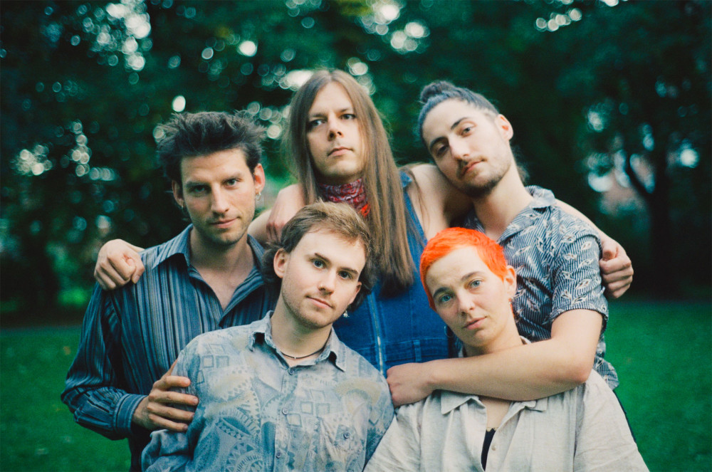 Man sieht fünf Personen auf einer Wiese, zwei im Vordergrund und dreibi Hintergrund. Alle haben die Arme um ihre nebenstehende Person gelegt. Alle lächeln ein wenig und schauen ruhig in die Kamera