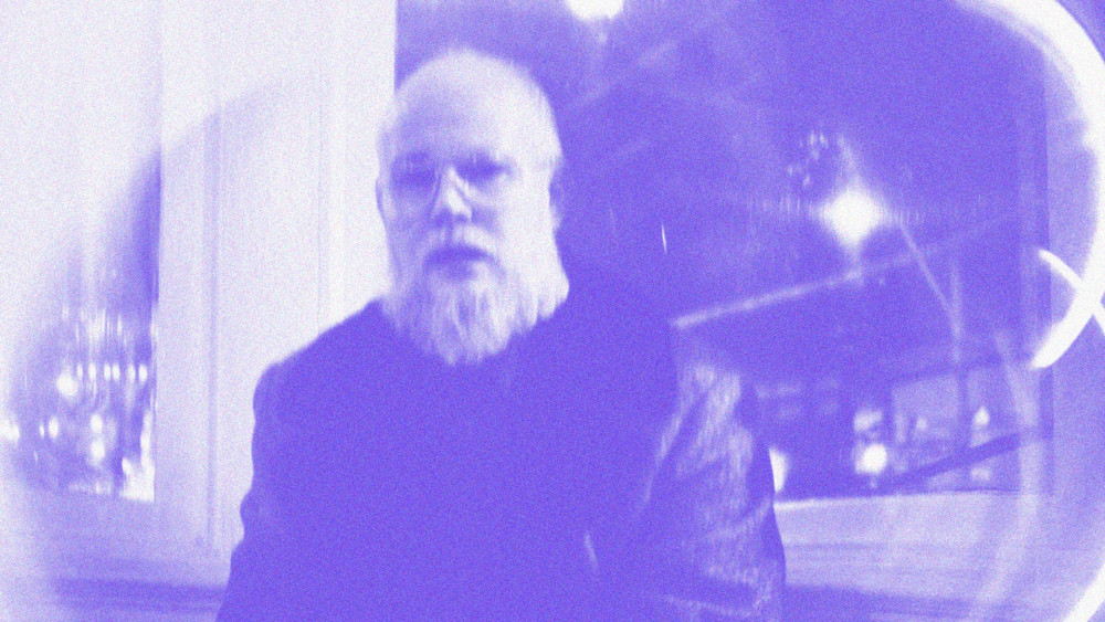 Auf dem Bild sieht man eine männlich gelesene Person. Das Bild ist etwas verschwommen. Die Person hat fast eine Glatze, trägt eine Brille und hat einen langen Bart. Außerdem trägt Die Person eine schwarze Lederjacke.