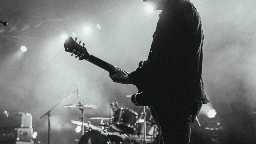 Auf dem Bild ist eine Person auf einer Bühne zu sehen. Die Person ist von hinten seitlich zu sehen und speilt auf einer Gitarre. Im Hintergrund sieht man ein Schlagzeug und oben an der Decke Lampen. Der Kopf der Person ist nicht zu sehen.