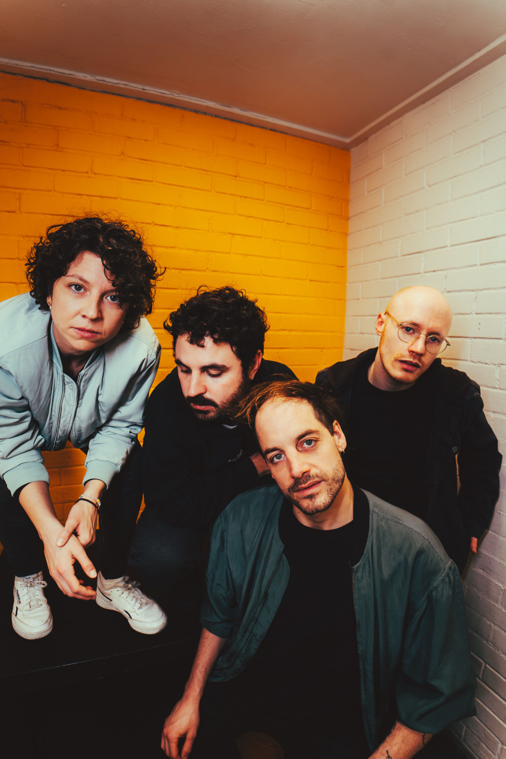 Es sind 4 Personen zu sehen vor einer gelben und einer weißen Steinwand. drei von ihnen sind männlich gelesen, von denen zwei auch in die Kamera schauen. die Vierte Person ist weiblich gelesen und steht während sie sich runter beugt um ebenfalls in die Kamera zu schauen.