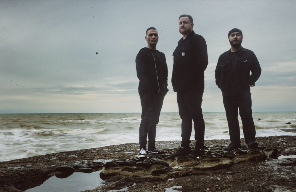 Auf dem Bild sind drei männlich gelesene Personen am Meer zu sehen. Alle Personen trage schwarze Klamotten. Sieh stehen am Strand und im Hintergrund ist Meer zu sehen. Es ist eher schlechtes Wetter.