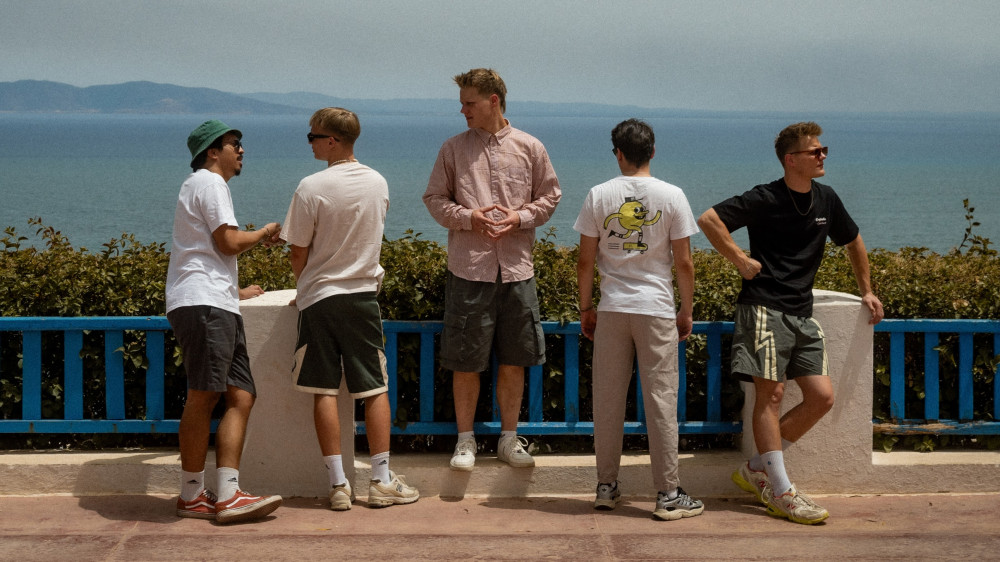 Auf dem Bild sind fünf männlich gelesene Personen zu sehen. Im Hintergrund sieht man das Meer. Die Person in der Mitte und ganz rechts schauen nach vorne, die anderen aufs Meer. Die Person ganz links trägt einen Hut. Alle haben kurze Haare