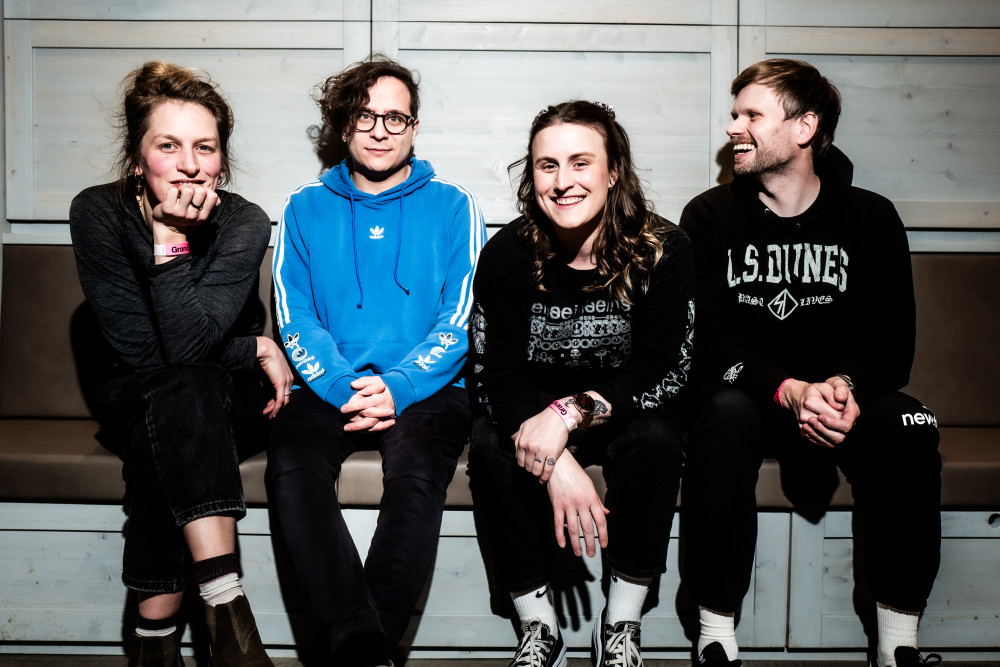 Auf dem Bild sind 4 Personen zu sehen. 2 weiblichgelesene Personen und eine männlichgelesen Person, die alle jeweils schawarz tragen. die vierte Person trägt einen Blauen Pullover. Sie sitzen alle auf einer Bank mit weißem Holz Hintergrund.