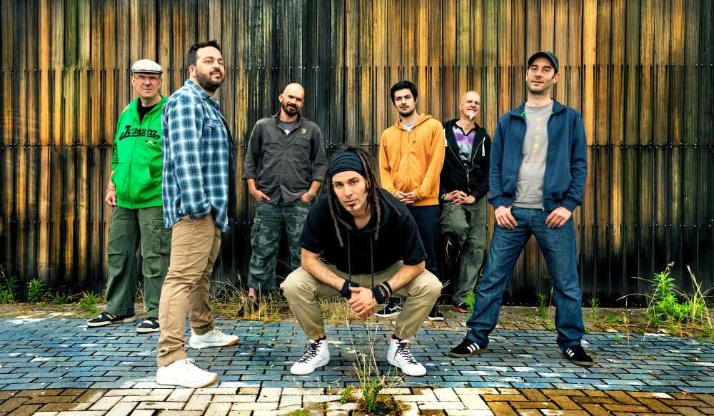 Auf dem sind acht männlich gelesene Personen zu sehen. Alle befinden sich vor einer Holzwand. Die person vorne in der Mitte sitzt in der Hocke und jeweils eine Person steht rechts, eine links. Die anderen vier Personen stehen in zweiter Reihe. Alle schauen freundlich.