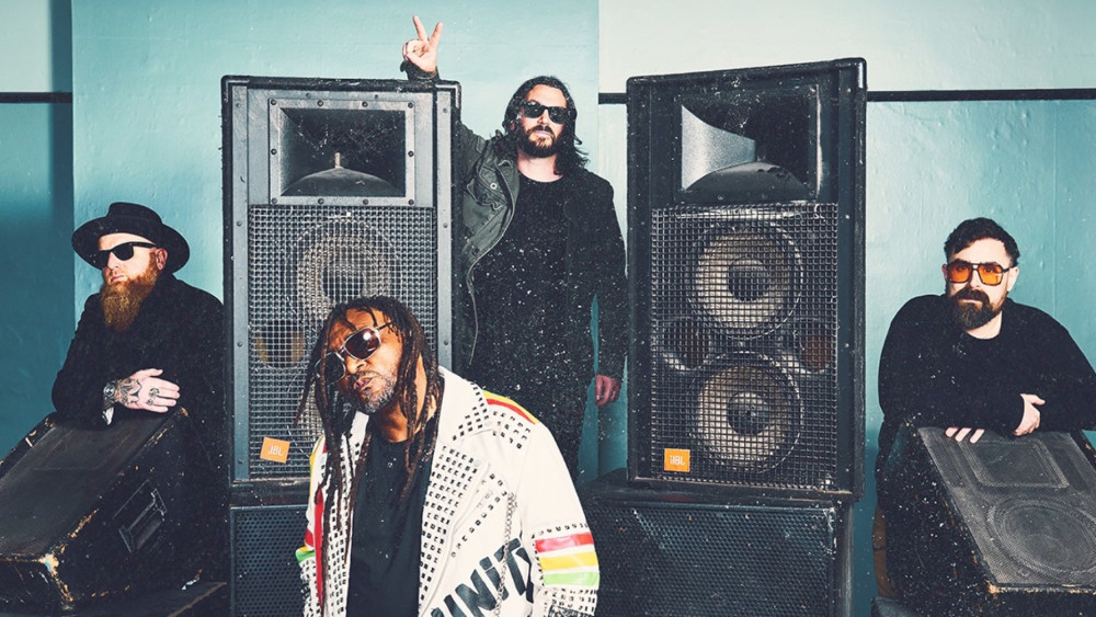 Auf dem Bild sind vier männlich gelesene Personen zu sehen. Die eine Person steht hinten zwischen zwei großen Boxen und hat den rechten Arm auf der Box abgestürzt und zeigt das Victory Zeichen. Die Person hat lange Haare und trägt wie alle anderen auf dem Bild eine Sonnenbrille. Die beiden Personen ganz außen sitzen und haben eine Box halb auf dem Schoß. Die Person ganz links hat einen lange Bart. Die Person vorne in der Mitte hat lange Harre und den Kopf etwas zur Seite gelegt.