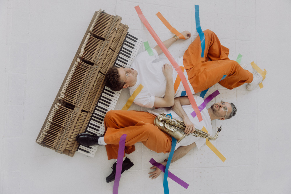 Auf dem Foto sind zwei Personen auf dem Boden liegend zu sehen. Die eine Person liegt mit dem Kopf auf der Kalviertastatur die zu sehen ist, hat kurze Haare und trägt eine Brille. Die andere Person liegt daneben. Allerdings mit dem Kopf nach unten und dem linken Fuß aufgestellt auf der Klaviertastatur. Auf dem Oberkörper der Person liegt ein Saxophon, welches die Person mit der rechten Hand festhält. Die Person hat ebenfalls kurze Haare und trägt eine Brille.