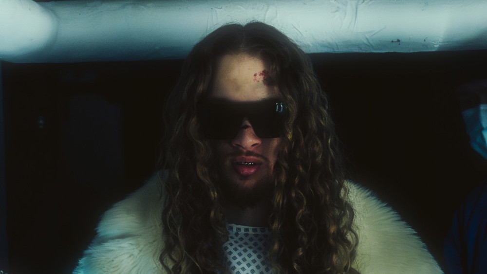 Auf dem Bild ist eine männlich gelesene Person zu sehen. Die Person hat lange lockige Haare, einen Bart und trägt eine Sonnenbrille. Die Person trägt eine art weißen Fellmantel.