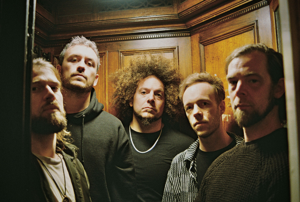 Auf dem Bild sind fünf männlich gelesene Personen zu sehen in dunkler Kleidung, die alle in einem Aufzug stehen.