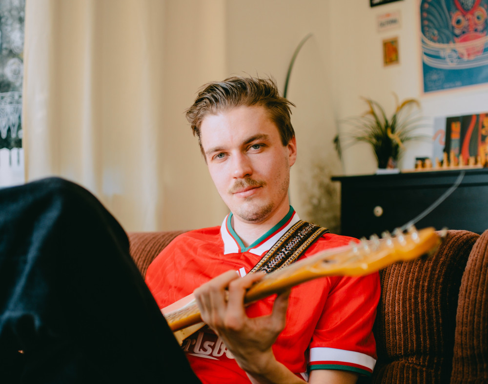 Auf dem Bild ist eine männlich gelesene Person zu sehen, welche auf einem braunen Sofa sitzt und Gitarre spielt. Diese trägt ein rotes Trikot, und eine schwarze Hose.