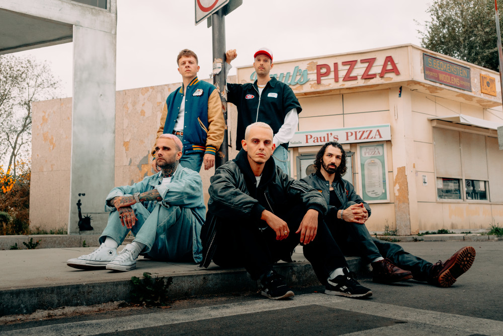 Auf dem Bild sind fünf männlich gelesene Personen zu sehen. Sie befinden sich vor einer Pizzeria. Drei Personen sitzen vorne, die anderen beiden stehen hinten. Die Personen vorne links und in der Mitte haben beide kurze Haare, die Person vorne rechts hat lange lockige Haare. Die beiden Personen hinten tragen College Jacken. Alle schauen neutral.