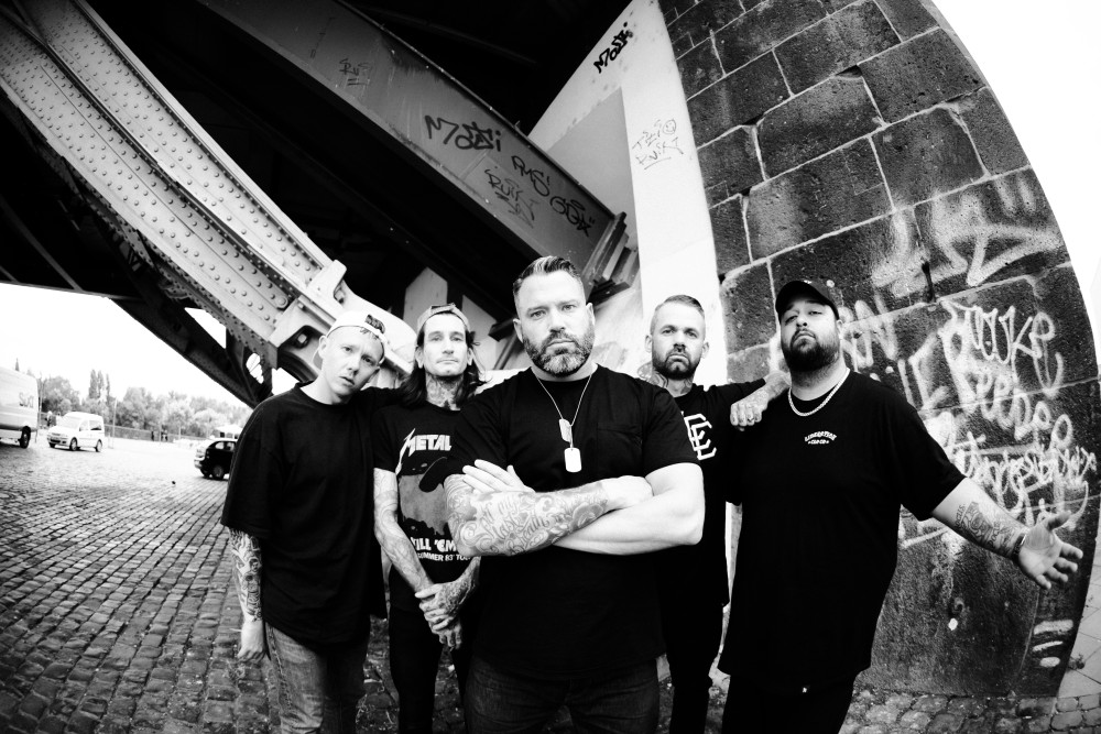 Auf dem Bild sind fünf männlich gelesene Personen sehen, welche alle frontal und ernst in die Kamera schauen. Alle sind schwarz gekleidet.