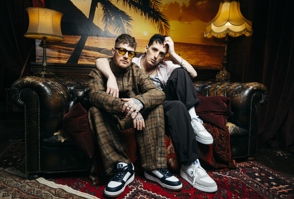 Auf dem Bild sitzen zwei männlich gelesene Personen auf einer Couch. Links und rechts auf der Couch befindet befindet sich jeweils eine Lampe. Hinter der Couch an der Wand hängt ein Bilderrahmen.