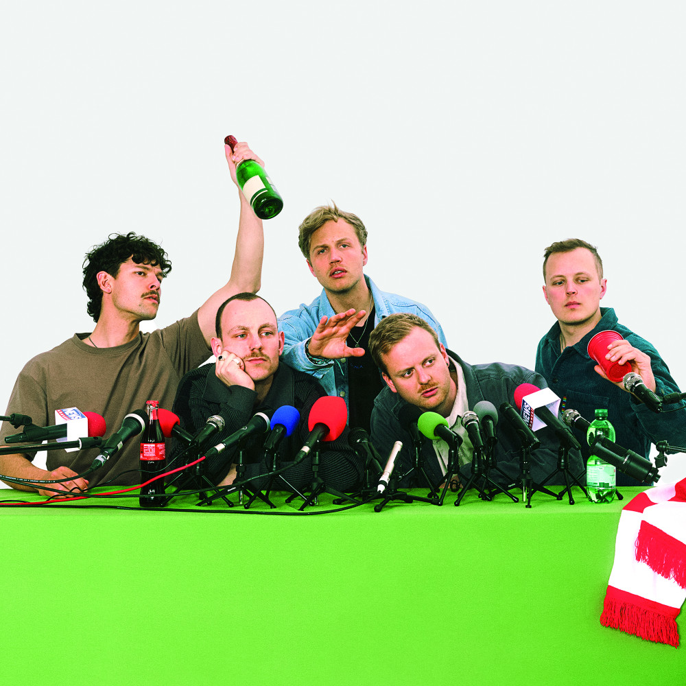 Auf dem Bild sind fünf männlich gelesene Personen zu sehen. Sie sitzen in einem Pressekonferenz Setup. Vier sitzen in einer Reihe, eine Person ist hinter den anderen.